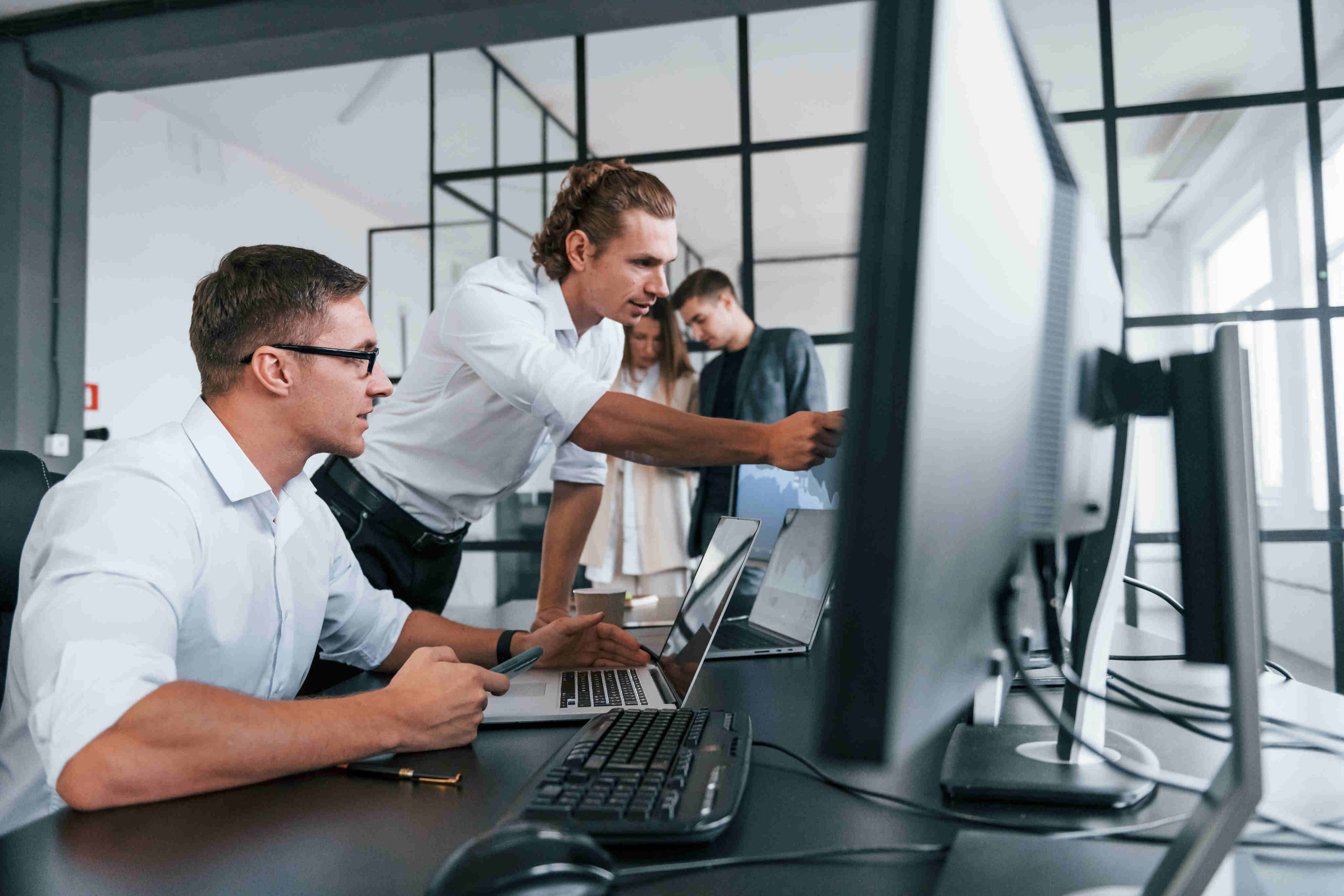 Programátoři společně řeší úkol u počítače v moderní kanceláři. Jeden z nich ukazuje kolegovi důležitý detail